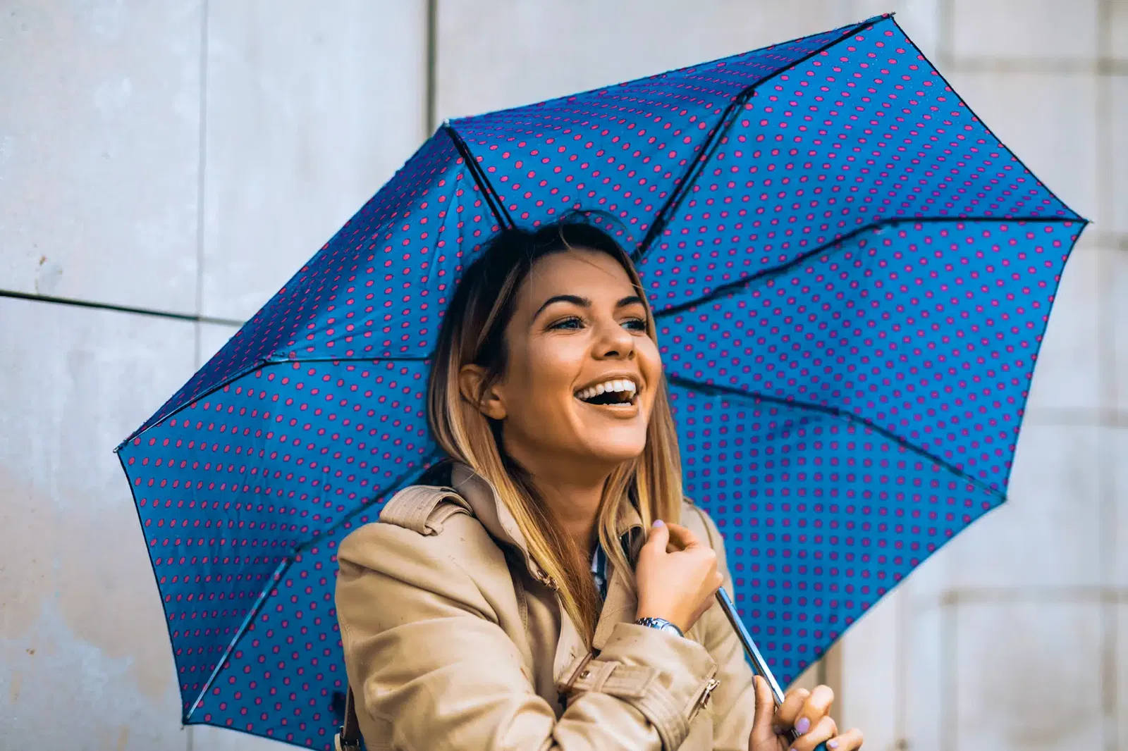 A woman in a beige coat smiles while holding a blue and pink polka-dotted umbrella, knowing her emergency fund gives her peace of mind no matter the weather.