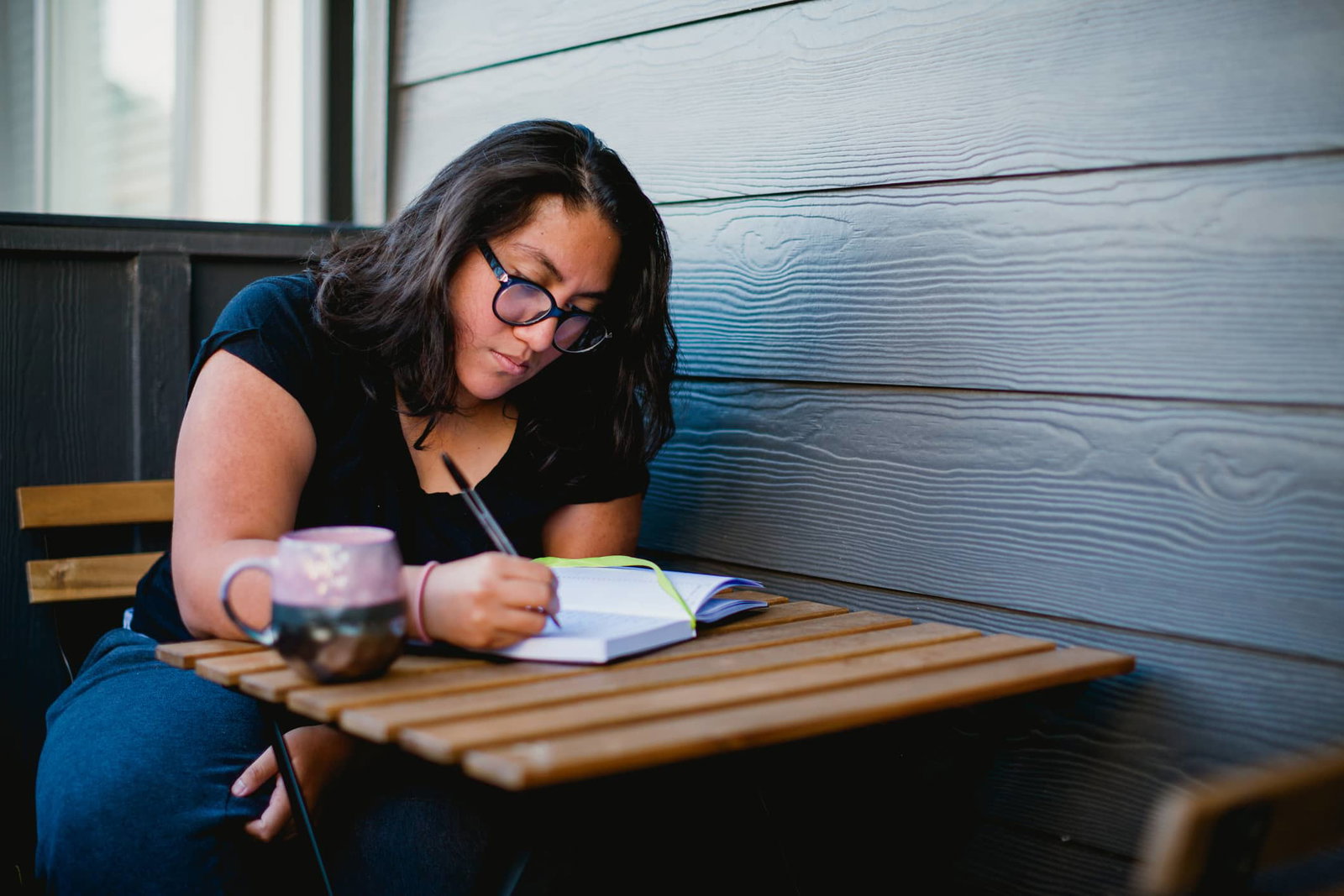 A person with glasses writes in a notebook while sitting at a wooden table with a mug beside them.