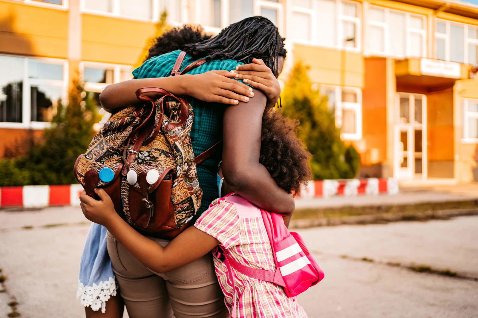 An adult with a patterned backpack hugs three children outside a school building. The children also carry backpacks.