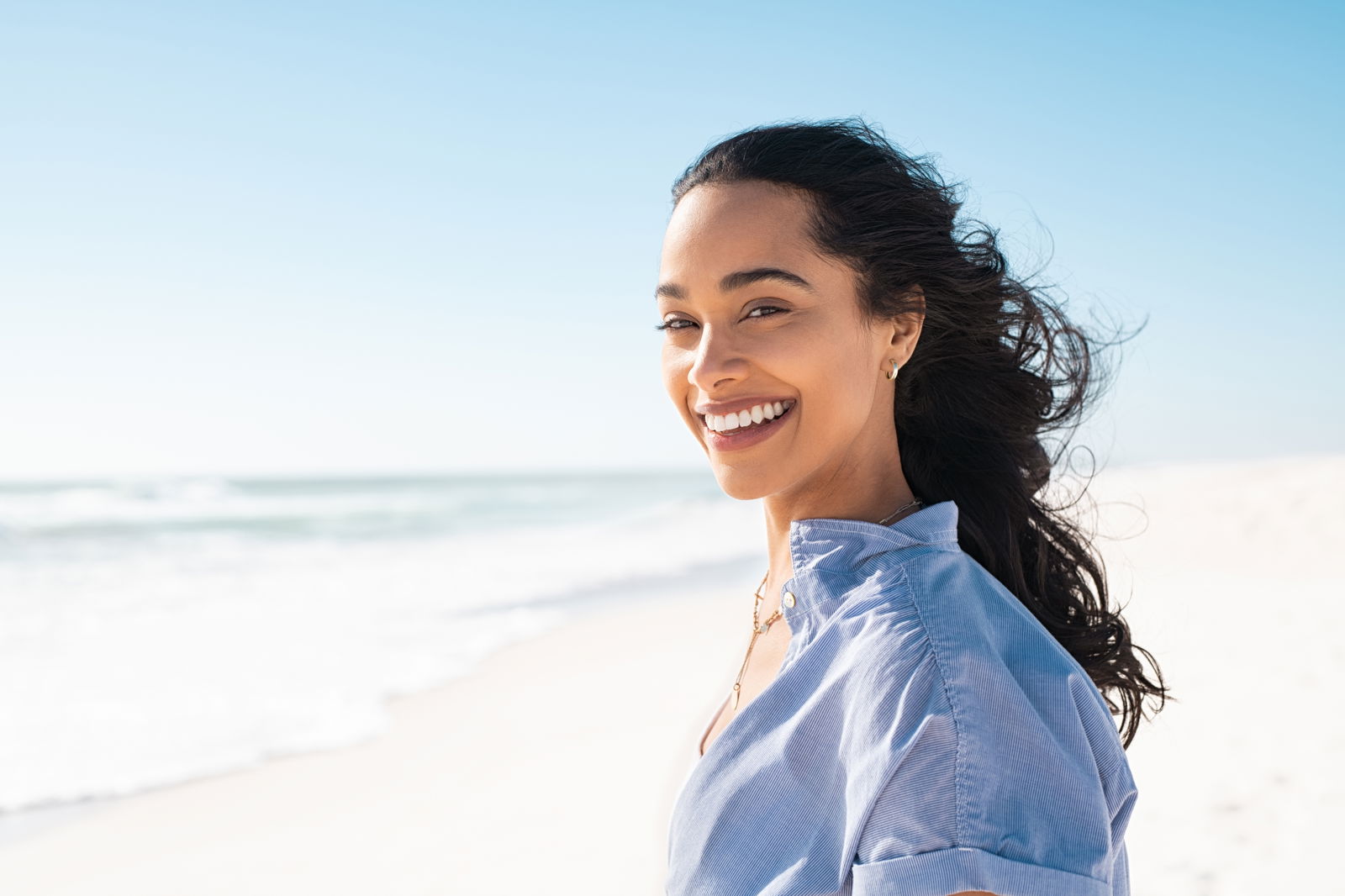 A smiling woman with long hair stands on a beach, wearing a blue shirt, with the ocean and clear sky in the background.