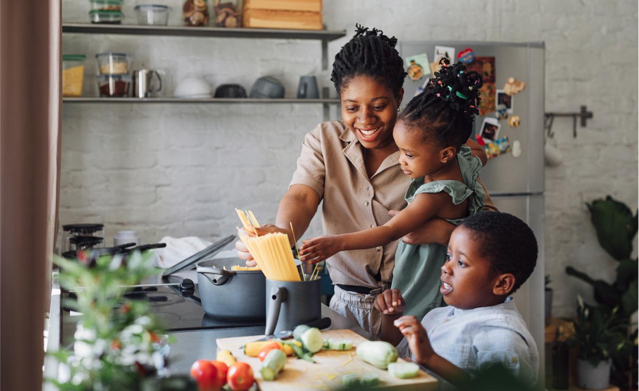 A woman, holding a girl, and a boy place uncooked spaghetti into a pot on the stove in a kitchen. Vegetables and a cutting board are on the counter nearby.