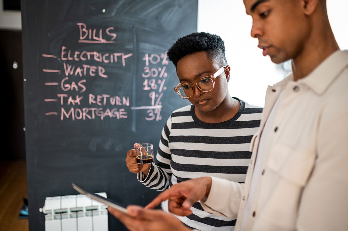 Two people review a tablet in front of a chalkboard listing bills with percentages for electricity, water, gas, tax return, and mortgage. One person holds a glass, and both appear focused.