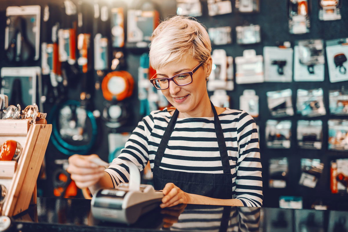 A person in a striped shirt and apron processes a payment at a retail store counter surrounded by various products.