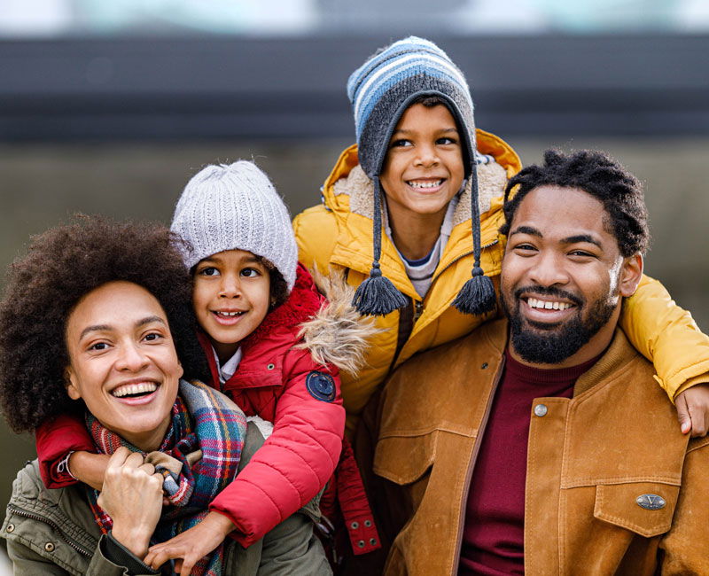 A smiling family of four wearing winter coats and hats stands outdoors, with one child on the father's shoulders.