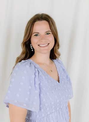 Woman with long brown hair wearing a light purple dress stands in front of a plain white background, smiling at the camera.