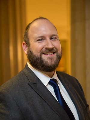 A man with a beard and short hair, wearing a grey suit, white shirt, and navy tie, smiles at the camera in an indoor setting with warm lighting.