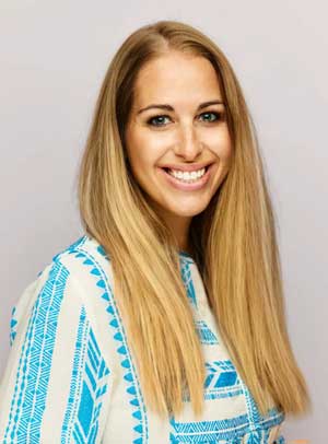 A woman with long straight blonde hair wearing a white shirt with blue geometric patterns smiles at the camera against a plain light-colored background.