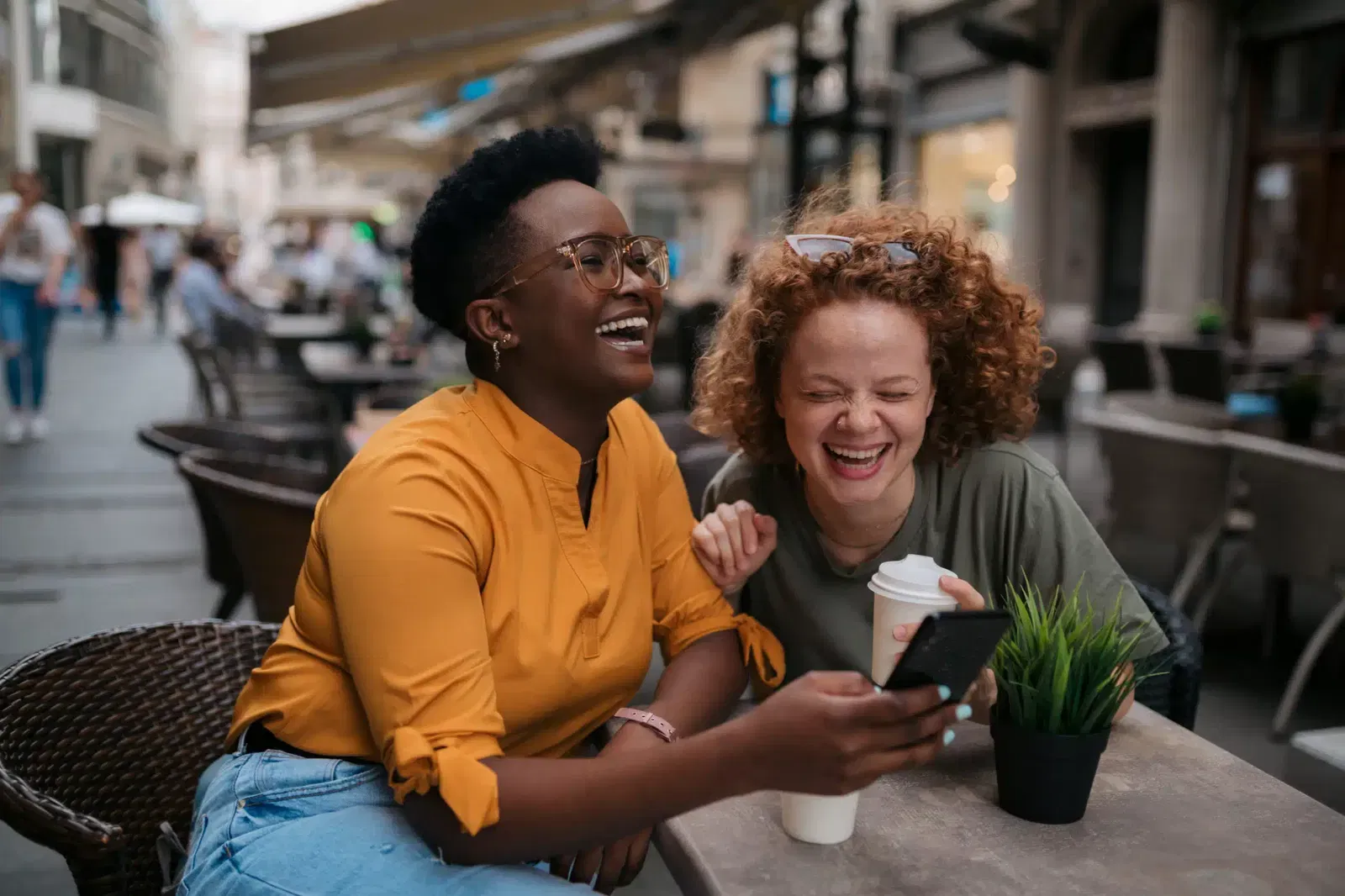 Two women sit at an outdoor café table, laughing together. One holds a smartphone and a coffee cup, while the other also holds a coffee cup. There is a small plant on the table.
