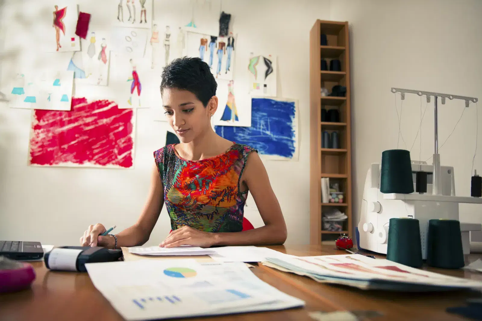A woman sits at a desk working with documents and charts, surrounded by fashion sketches and a sewing machine in a workspace.