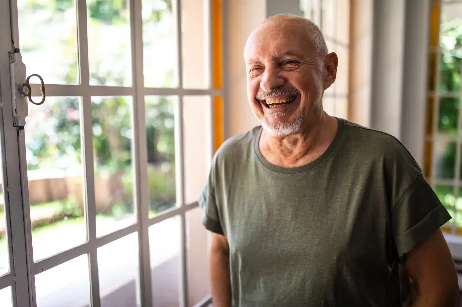 An older man with a short beard and gray hair smiles while standing indoors near large windows with sunlight streaming in.